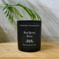 A black scented candle jar labeled 'Heartman Fragrances Red Berry Rose' sitting on a light wooden surface with a white vase and green leaves in the background.