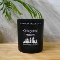 A black scented candle jar labeled 'Heartman Fragrances Cedarwood Amber' sits on a light wood surface with a soft neutral fabric backdrop and green leaves in the background.