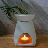 A white ceramic wax warmer with a lit tea light candle inside, set on a wooden surface against a textured gray wall with a green plant in the background.