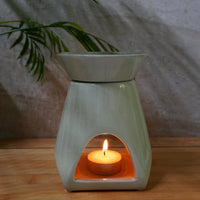 A ceramic wax warmer with a lit tea light candle inside, set on a wooden surface against a neutral textured background with a potted plant in the distance.