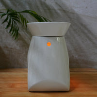 A white ceramic electric wax warmer with a small  tea light glowing on the front, set on a wooden surface against a textured gray wall with a plant in the background.