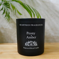 A black glass jar candle labeled 'Heartman Fragrances Peony Amber' sits on a light wooden surface with a soft beige fabric backdrop and green leaves in the background.