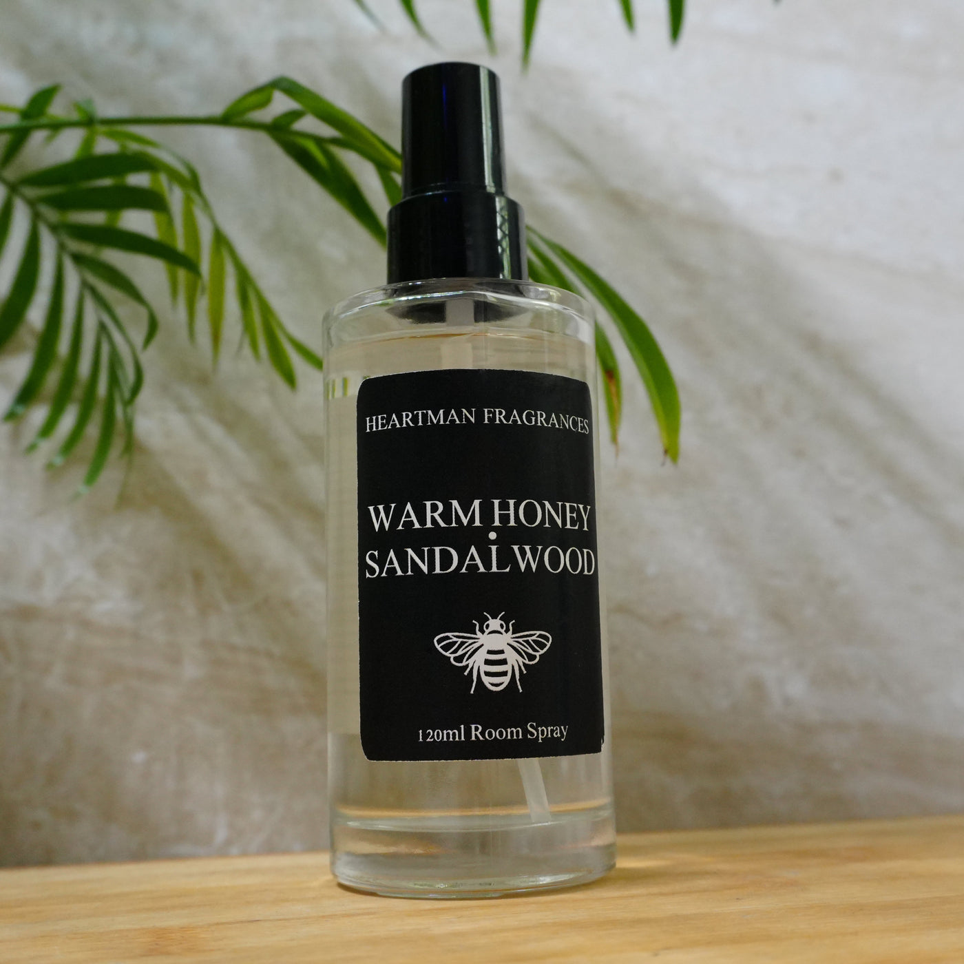 A 120ml room spray bottle of Heartman Fragrances 'Warm Honey Sandalwood' scent, featuring a black spray nozzle and a black label with white text, set on a light wooden surface with a textured beige wall and green leaves in the background.