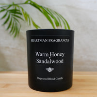 A black jar candle labeled 'Heartman Fragrances Warm Honey Sandalwood' sits on a light wood surface with a blurred indoor background and green plant leaves.