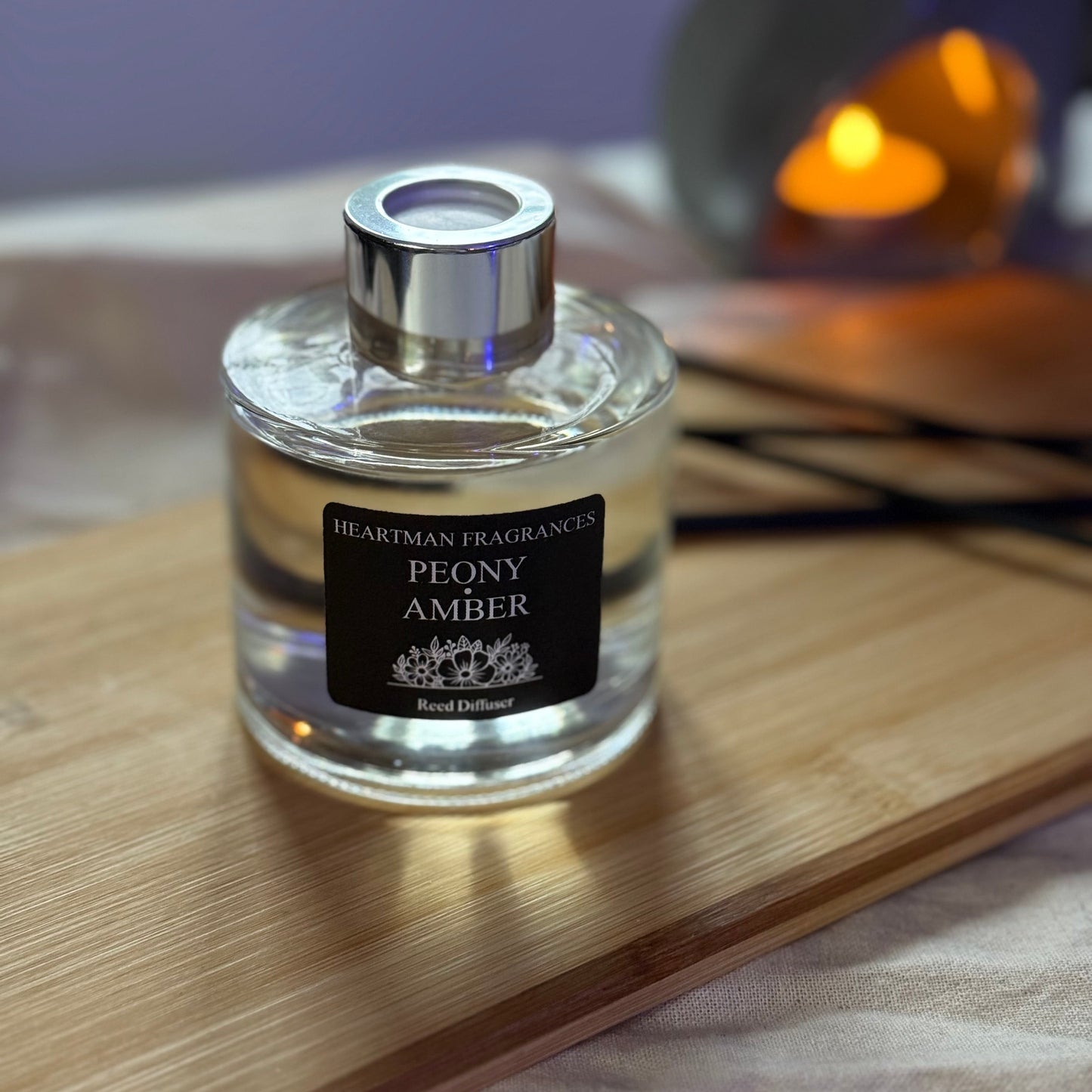 reed diffuser bottle labeled 'Peony Amber' by Heartman Fragrances on a wooden surface with a blurred candle in the background..