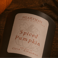 A close-up of a Heartman Autumn Collection candle labeled 'Spiced Pumpkin' with notes of clove and cinnamon, shown on a dark wooden surface with a pumpkin partially visible in the background.