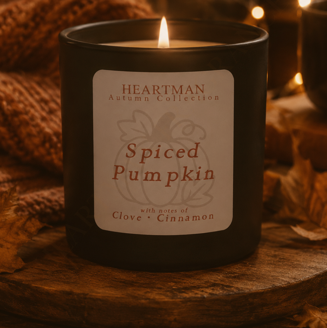 A lit Heartman Autumn Collection candle in a matte black jar, labeled 'Spiced Pumpkin' with notes of clove and cinnamon, set on a rustic wooden surface with a knitted blanket and warm bokeh lights in the background.